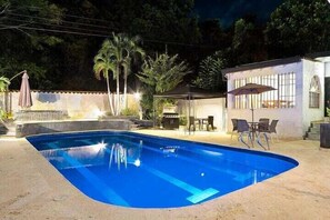 A swimming pool with a tiled deck, surrounded by palm trees and a patio area with outdoor furniture.