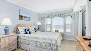 A bedroom with a bed, two nightstands, a dresser, a window with blinds, and a beach-themed wall art.