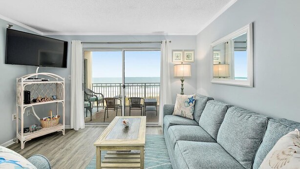 A living room with a grey sofa, a coffee table, a TV, a balcony with a view of the ocean, and a small table with chairs.