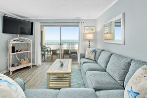 A living room with a grey sofa, a coffee table, a TV, a balcony with a view of the ocean, and a small table with chairs.