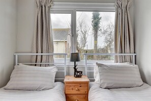 A bedroom with two beds, a wooden nightstand with drawers, a lamp, and a view of trees and a building through a window.