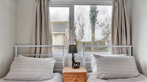Ein Schlafzimmer mit zwei Betten, einem Nachttisch aus Holz mit Schubladen, einer Lampe und Blick auf Bäume und ein Gebäude durch ein Fenster.