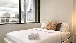 A neatly made bed with white bedding and rolled towels, a fluffy pillow, and a view of city buildings through a large window.