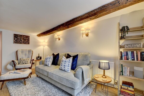 A cozy living room with a grey sofa, matching armchair, and a small round table with a lamp. A bookshelf filled with books is visible to the right.