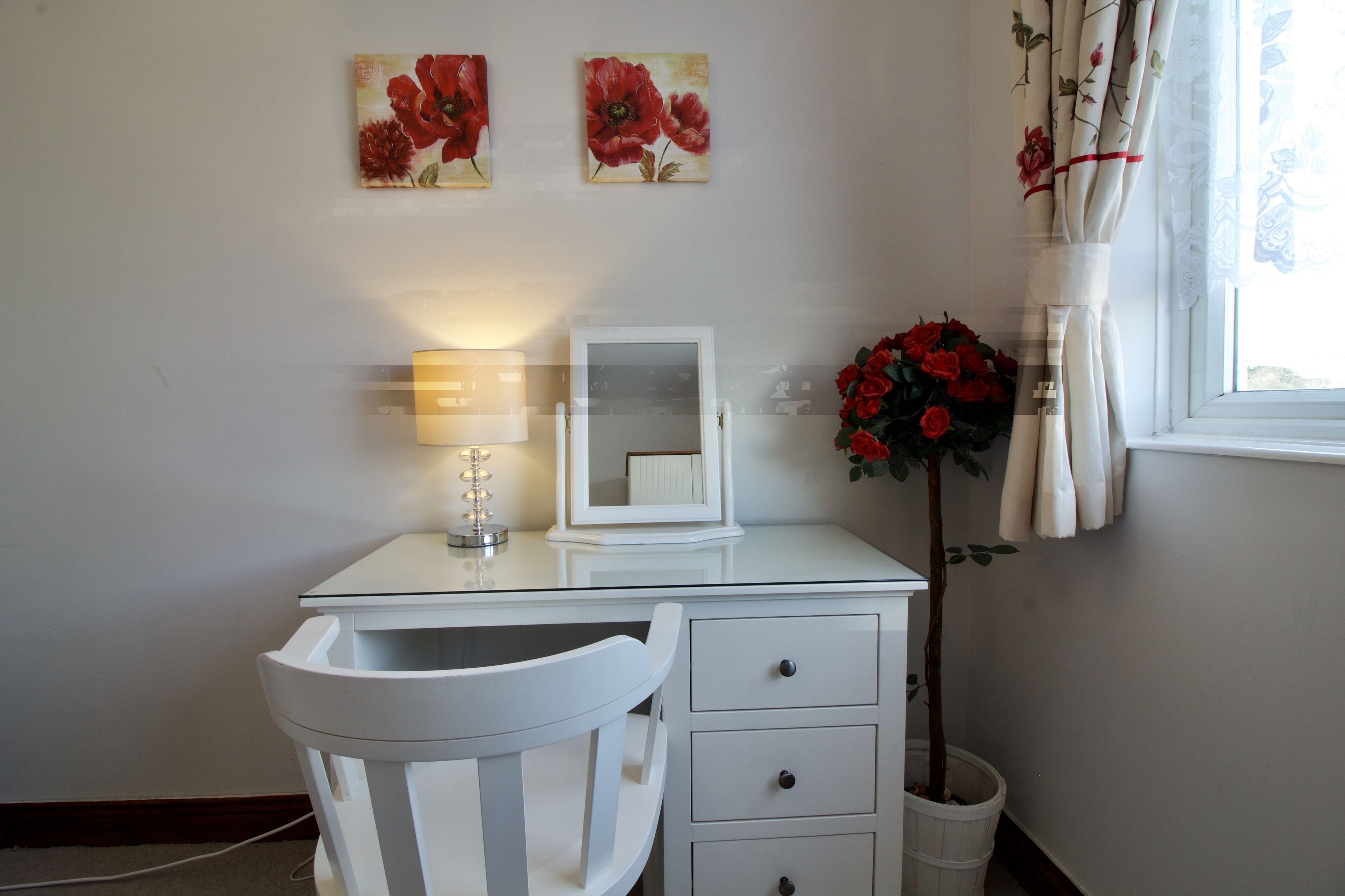 A white desk with a mirror, a lamp, and a chair. There are two paintings on the wall, a potted plant with red flowers, and a window with curtains.