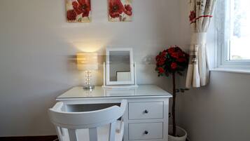A white desk with a mirror, a lamp, and a chair. There are two paintings on the wall, a potted plant with red flowers, and a window with curtains.