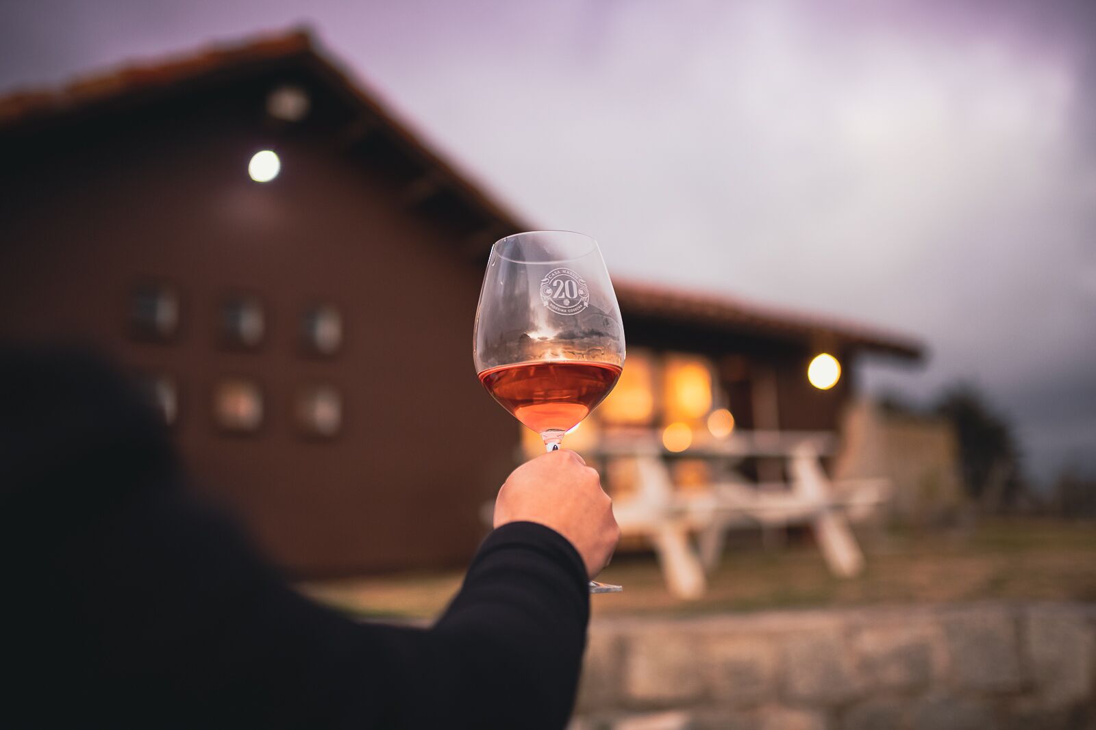 A person holding a wine glass with a logo, in front of a building with a wooden exterior and a lit interior.