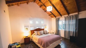 A bedroom with a wooden ceiling, a bed with a headboard, a nightstand, a chair, a wardrobe, and a window with curtains. - Villa Miramar at Casa Marin Vineyards (Cartagena)