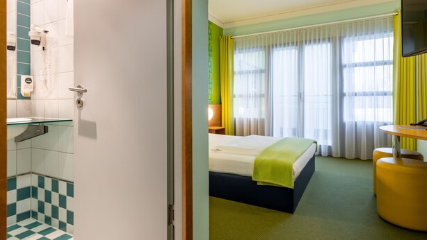 Una habitación de hotel con ducha, cama, mesita de noche, televisor y ventana con cortinas.