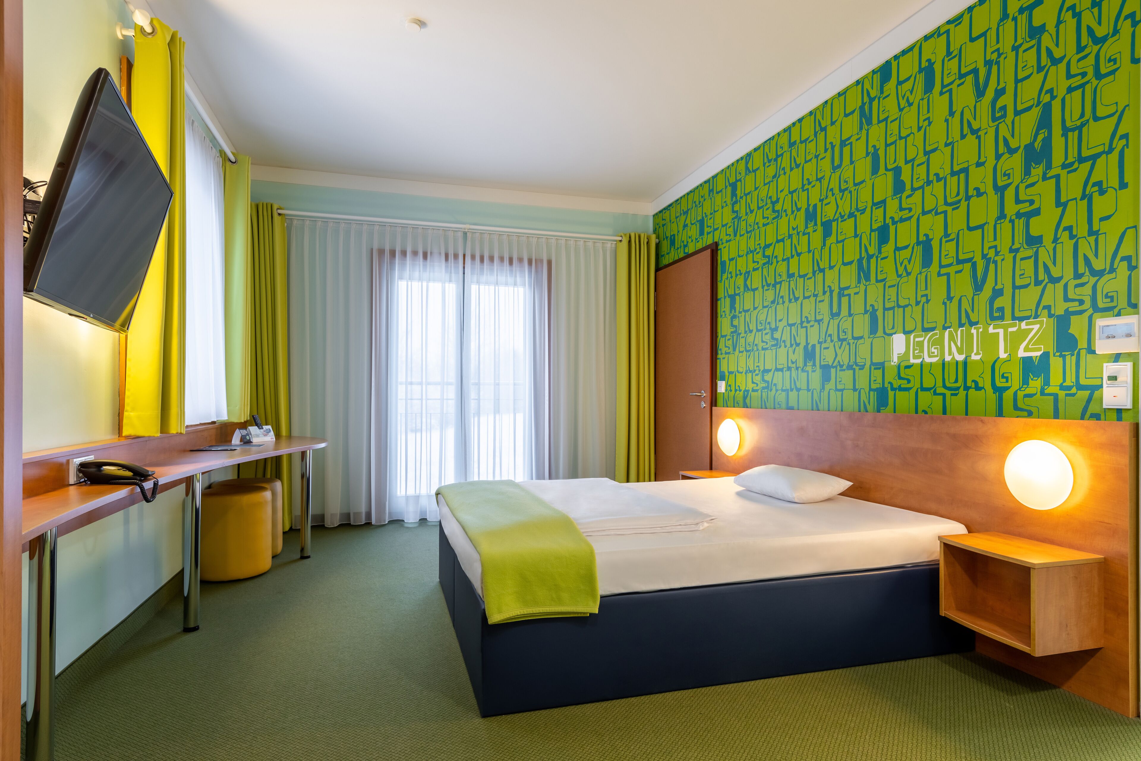 Una habitación de hotel con cama, escritorio, televisor y pared con estampado verde.