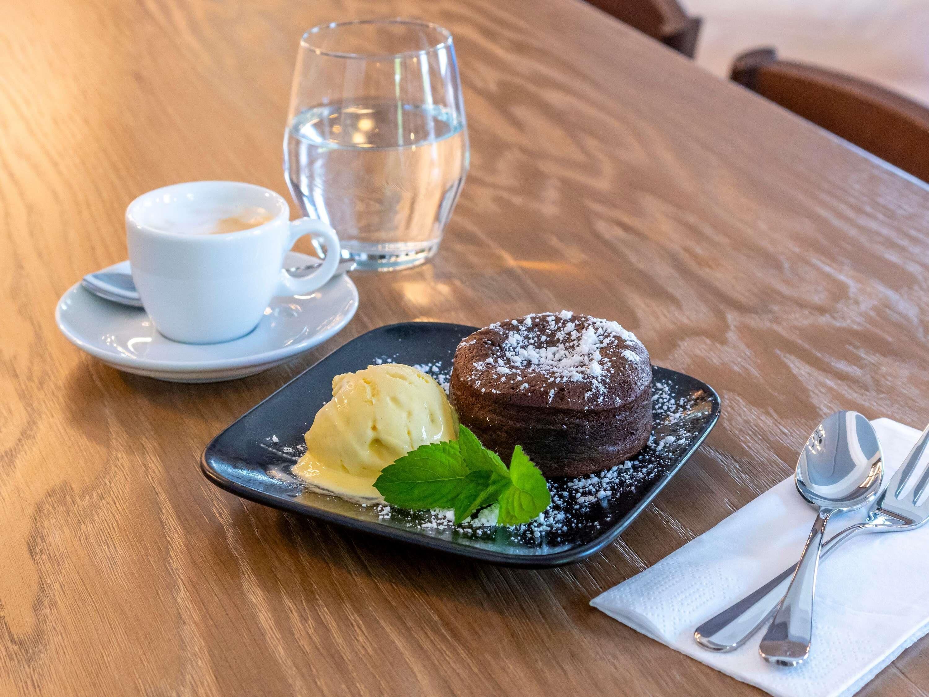 Un plato de postre con pastel de chocolate y helado, una taza de café y un vaso de agua sobre una mesa de madera.