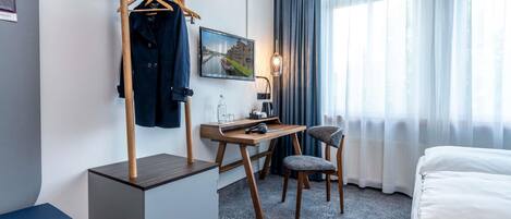Ett hotellrum med en säng, ett skrivbord med en stol, en garderob, en TV och ett fönster med gardiner.