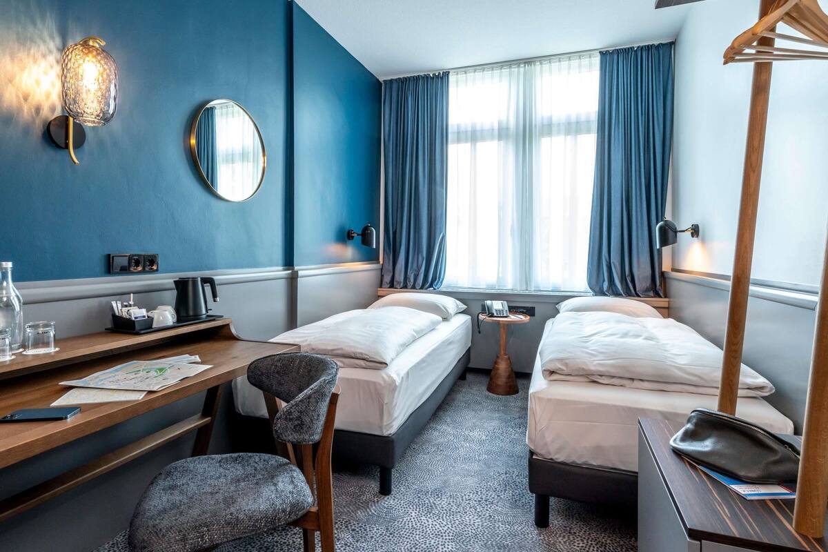 Habitación de hotel con dos camas, un escritorio con silla, un espejo, un teléfono y una ventana con cortinas.
