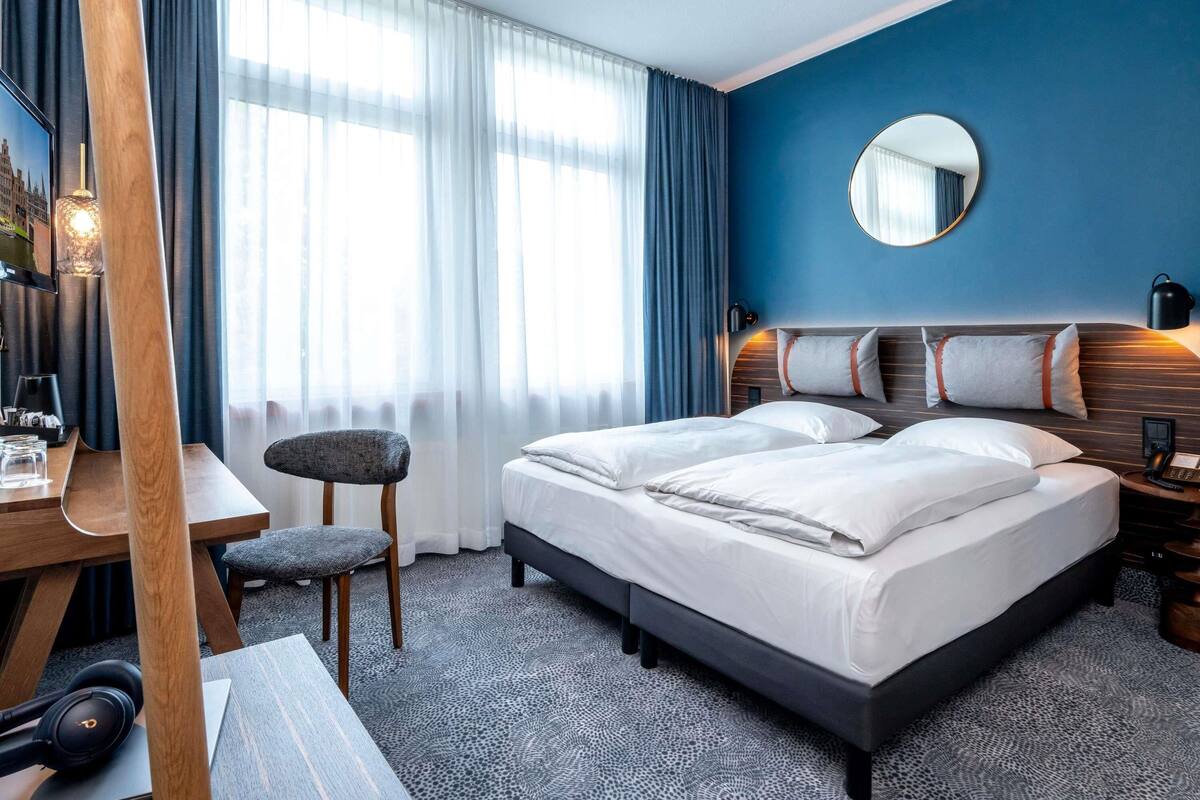 Habitación de hotel con una cama grande, un escritorio con una silla, un espejo redondo y una mesita de noche con lámpara.