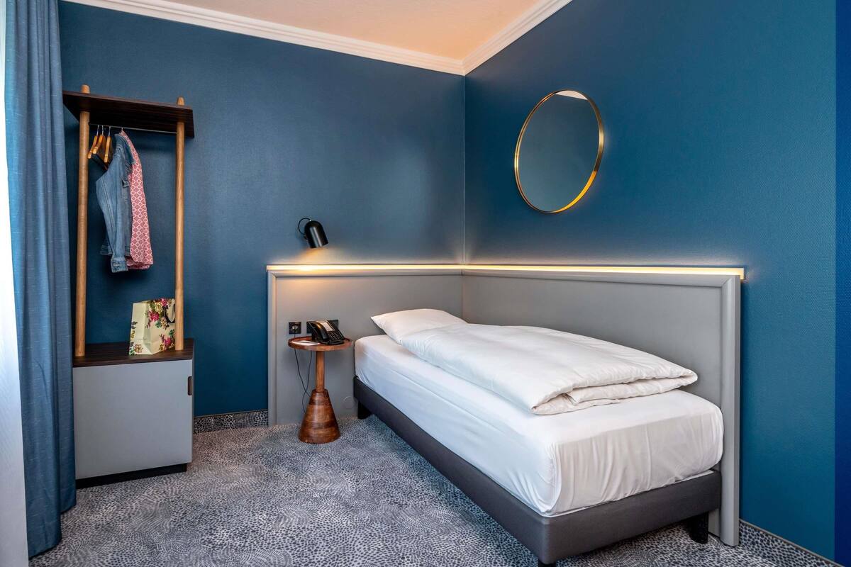 Una habitación de hotel moderna con una cama gris, una mesita de noche pequeña con una lámpara, un armario y un espejo redondo en la pared.