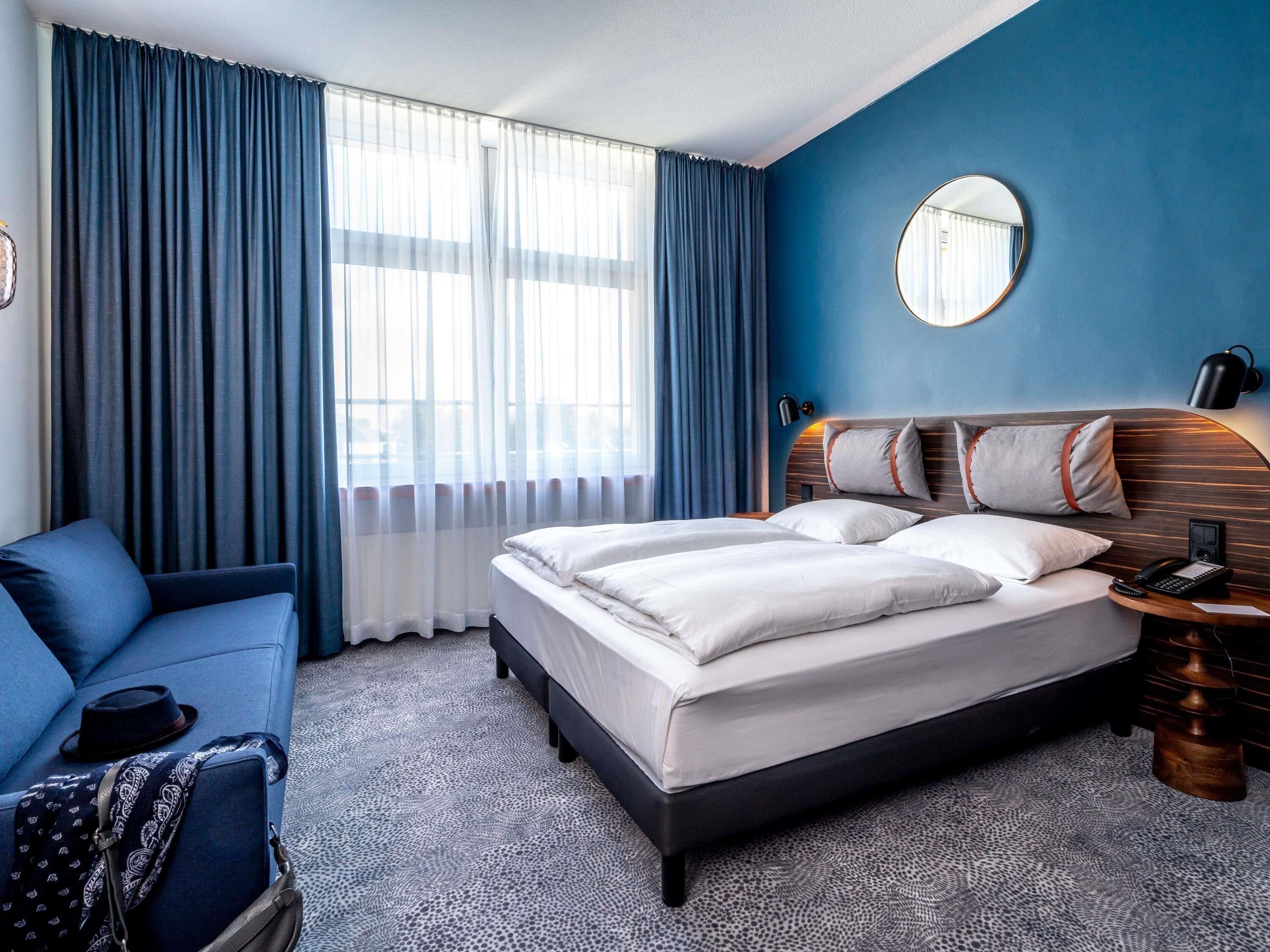 A hotel room with a bed, a blue sofa, a round mirror, and a nightstand with a lamp.