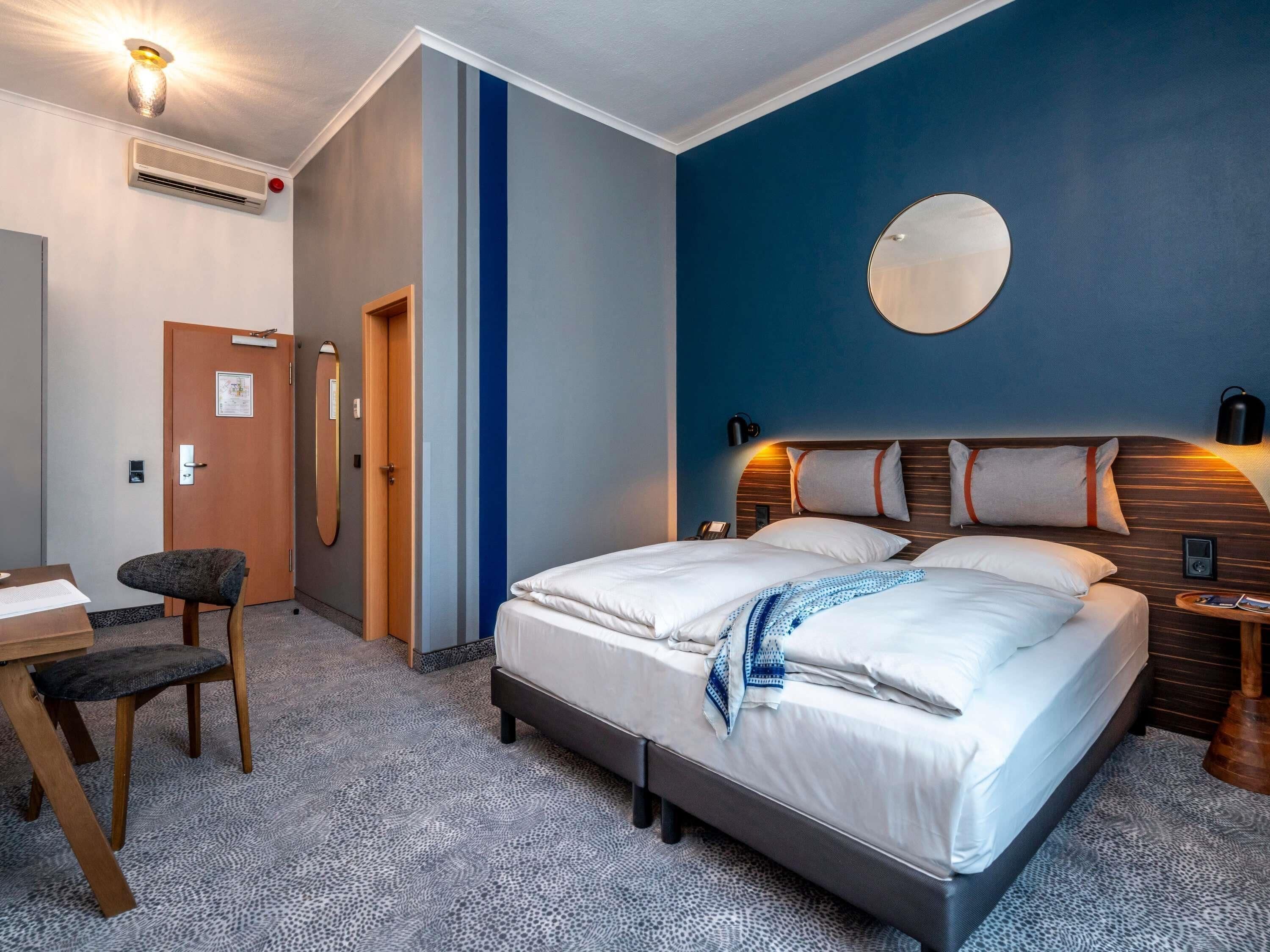A hotel room with a bed, a desk, a chair, a wall-mounted mirror, and a round mirror on the wall.