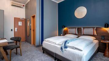 A hotel room with a bed, a desk, a chair, a wall-mounted mirror, and a round mirror on the wall.
