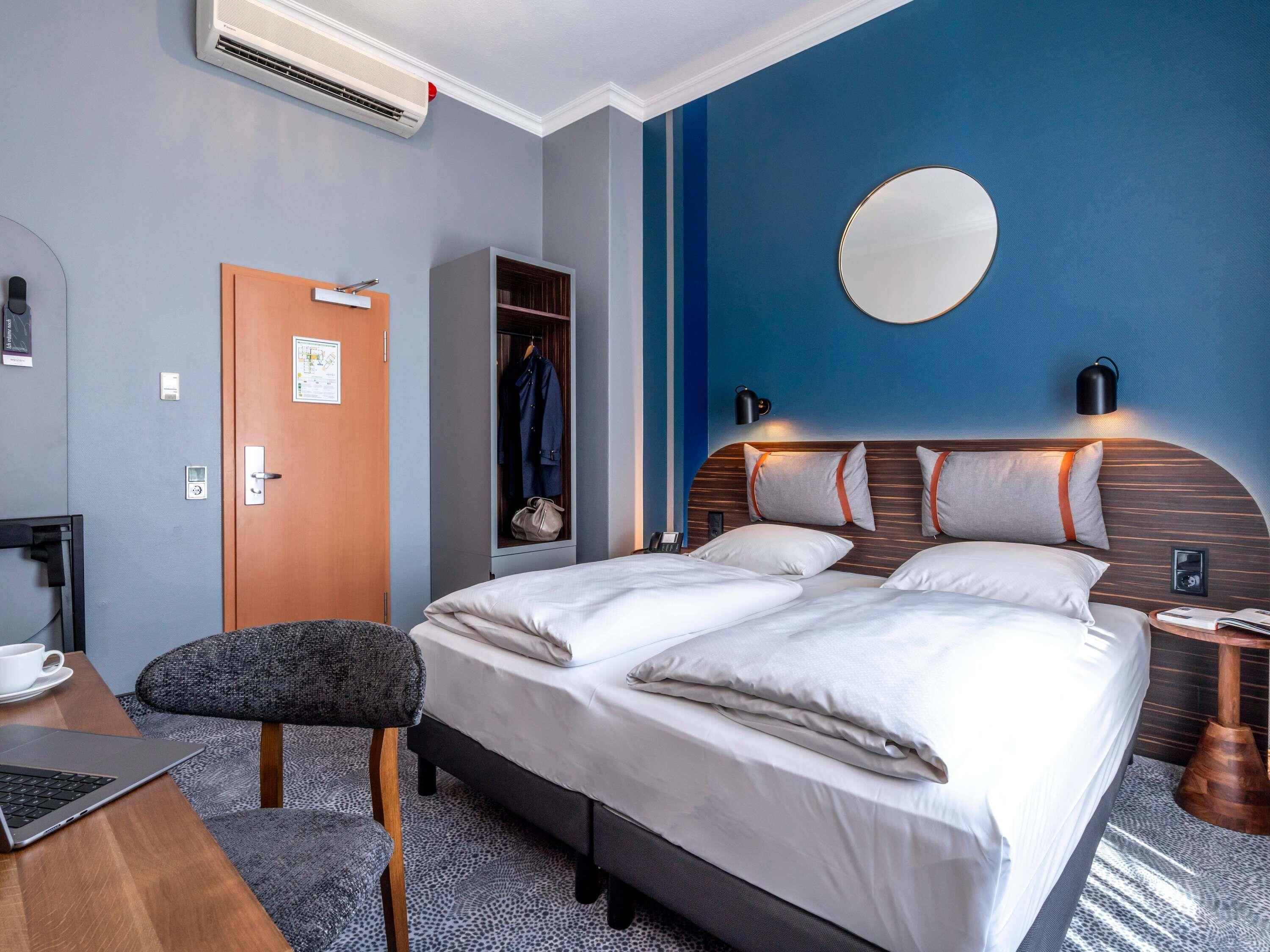 Ein modernes Hotelzimmer mit Bett, Schreibtisch mit Laptop, Kleiderschrank und einem wandmontierten Spiegel.