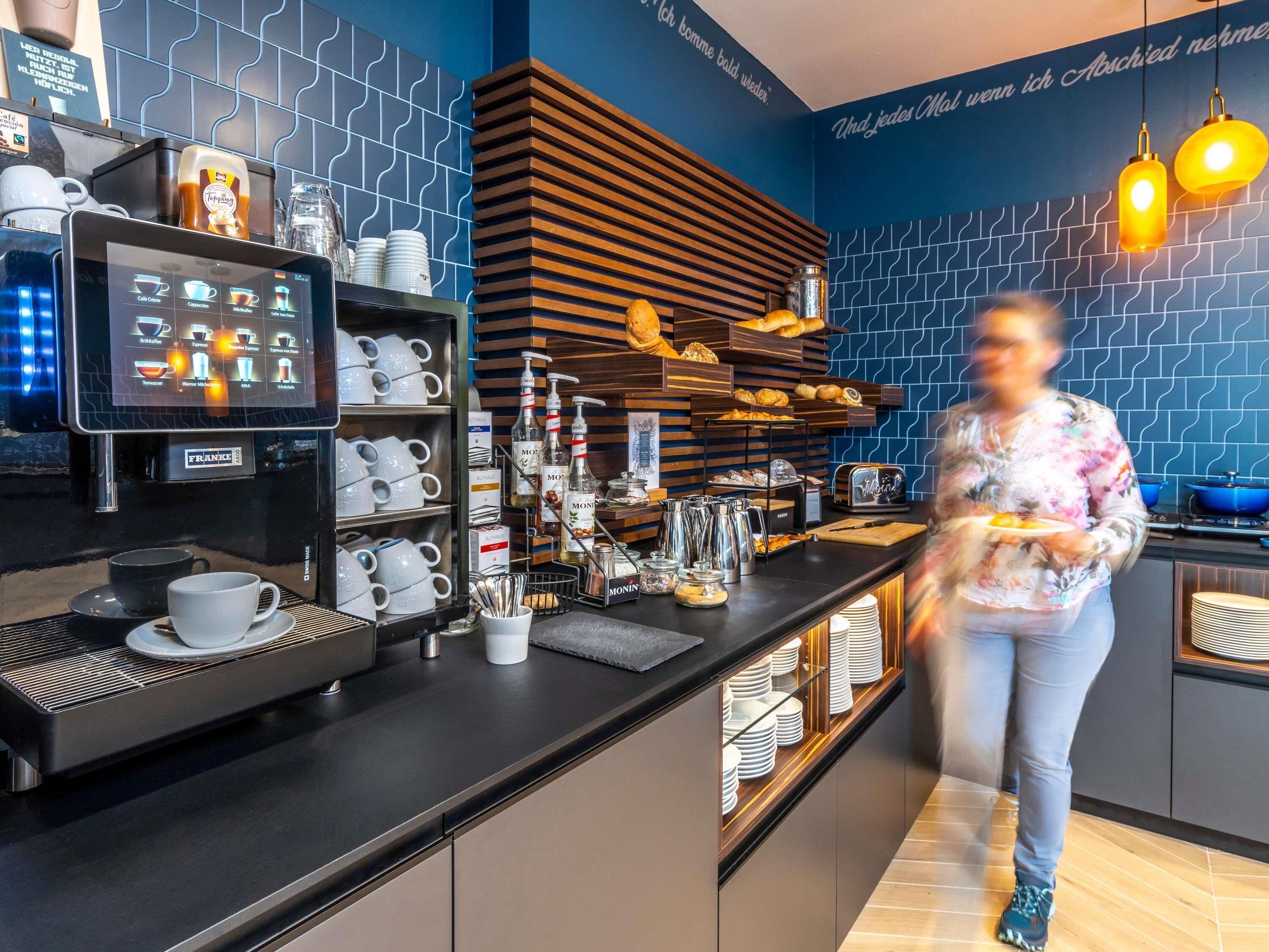 A modern coffee bar with a digital menu, coffee machines, and a variety of coffee cups and mugs.