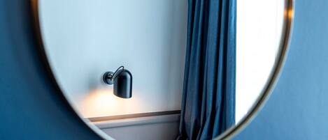 A round mirror reflecting a room corner with a wall-mounted light fixture and a window with curtains.