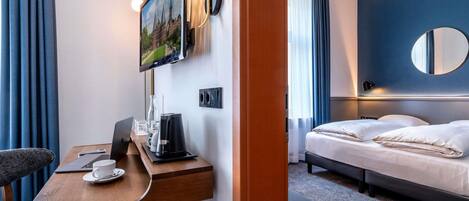Ein Hotelzimmer mit einem Bett, einem Schreibtisch mit Laptop, einer Kaffeemaschine und einem an der Wand befestigten Fernseher.