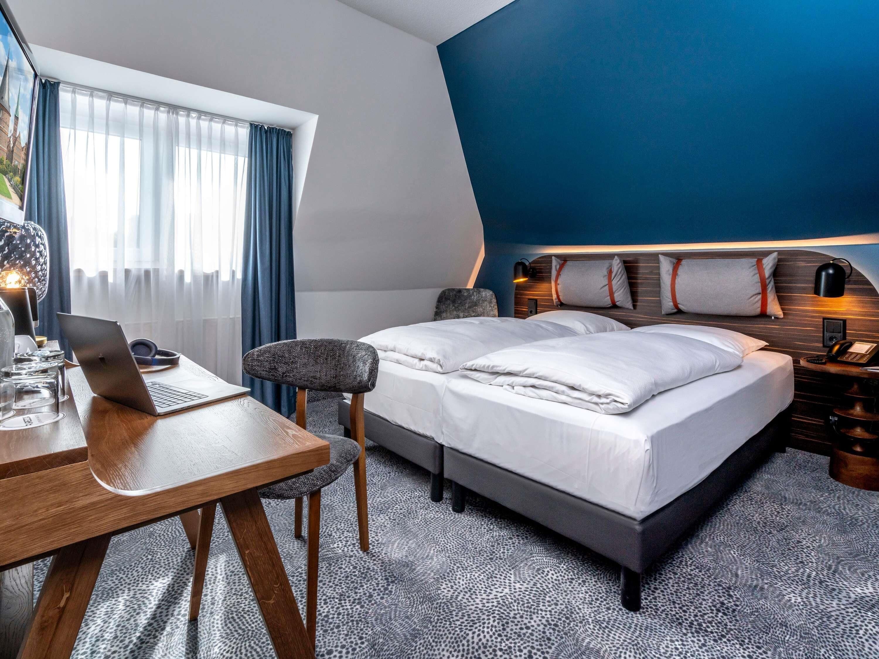 A hotel room with a bed, a desk with a laptop, a chair, a bedside table with a lamp, and a window with curtains.