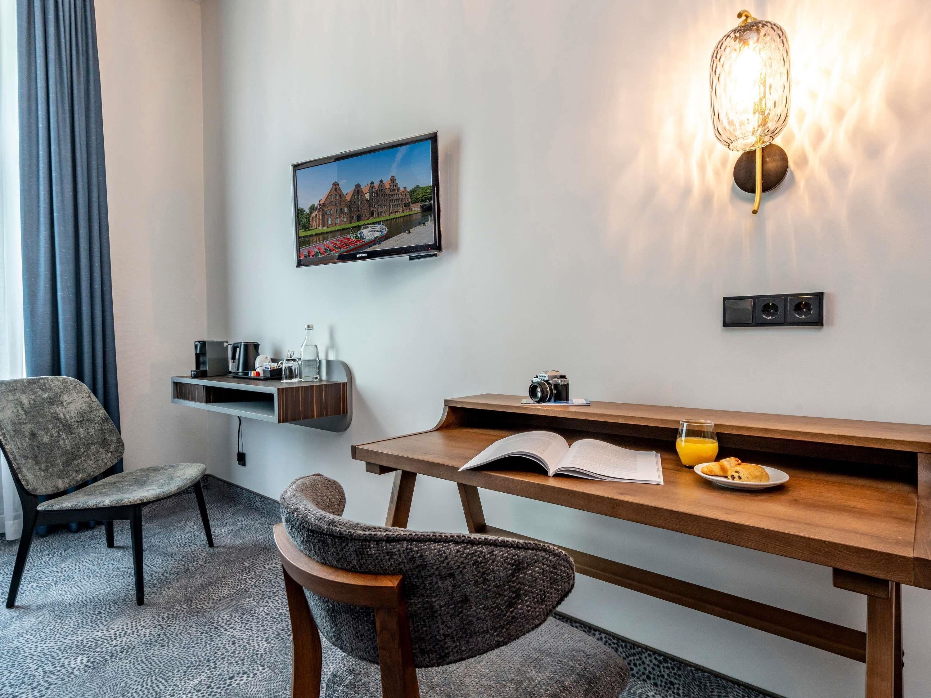 Ein Hotelzimmer mit einem Holztisch, einem Stuhl, einem an der Wand befestigten Fernseher, einer Wandleuchte und einem gerahmten Bild.