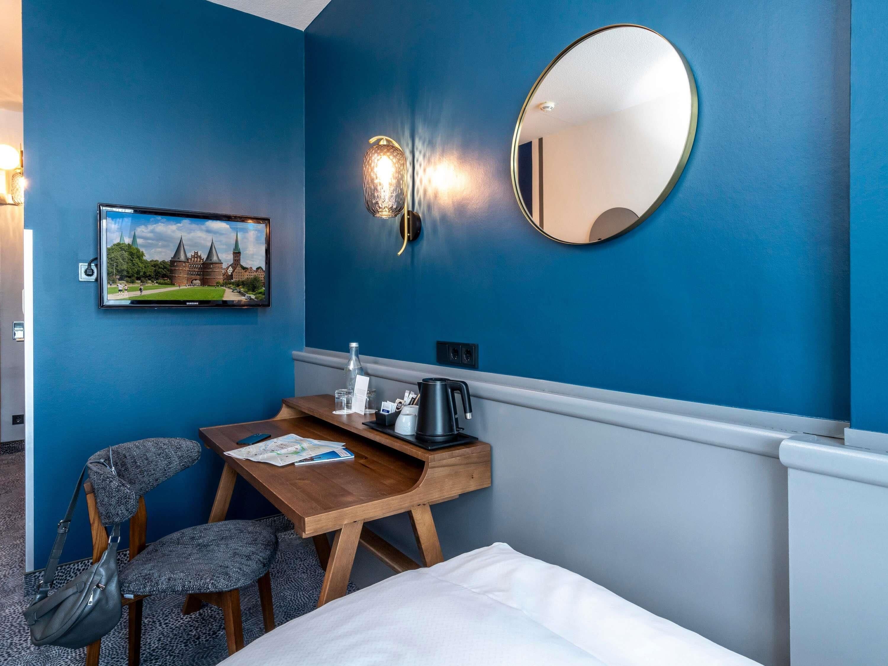 A hotel room with a bed, a desk with a kettle, a chair, a wall-mounted mirror, and a picture of a castle.