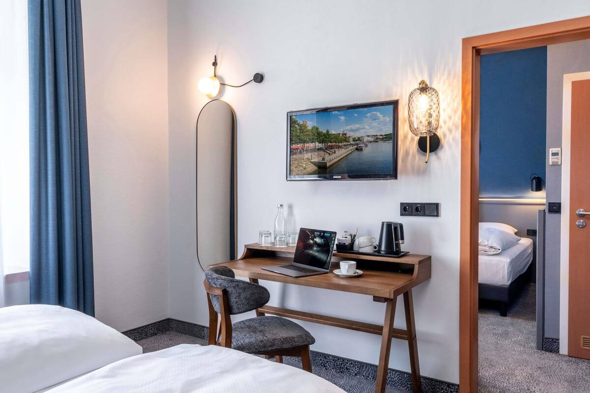 Una habitación de hotel con una cama, un escritorio con una computadora portátil, una silla, un televisor montado en la pared y una puerta que da a otra habitación.