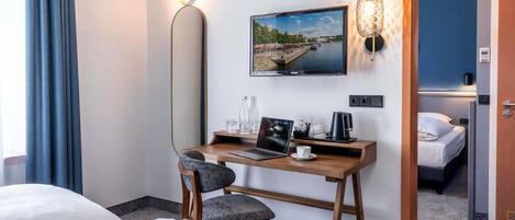 A hotel room with a bed, a desk with a laptop, a chair, a wall-mounted TV, and a door leading to another room.