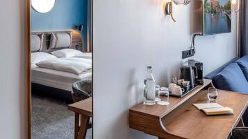 Ett hotellrum med en säng, ett skrivbord med en kaffebryggare, ett glas vatten och en tavla som föreställer en stadsbild.
