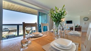 A dining area with a wooden table set for four, a vase of white flowers, and a view of the ocean through large windows. - 2 Bed in Westward Ho! (oc-seaspr) (Westward Ho!)