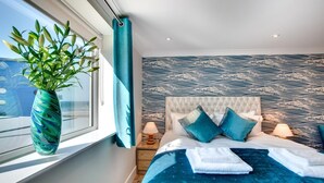 A bedroom with a large window, a plant, a bed with blue bedding, and a patterned wall. - 2 Bed in Westward Ho! (oc-seaspr) (Westward Ho!)