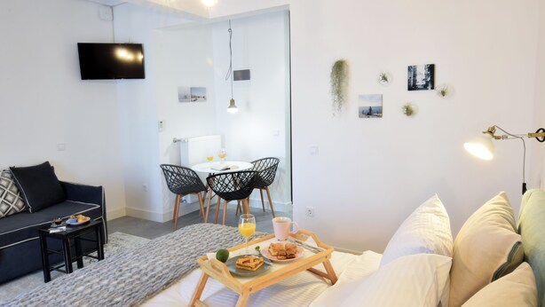 A modern living room with a sofa, a dining area with chairs, a TV mounted on the wall, and a breakfast tray on the bed. - Grey Studios (Thessaloniki)