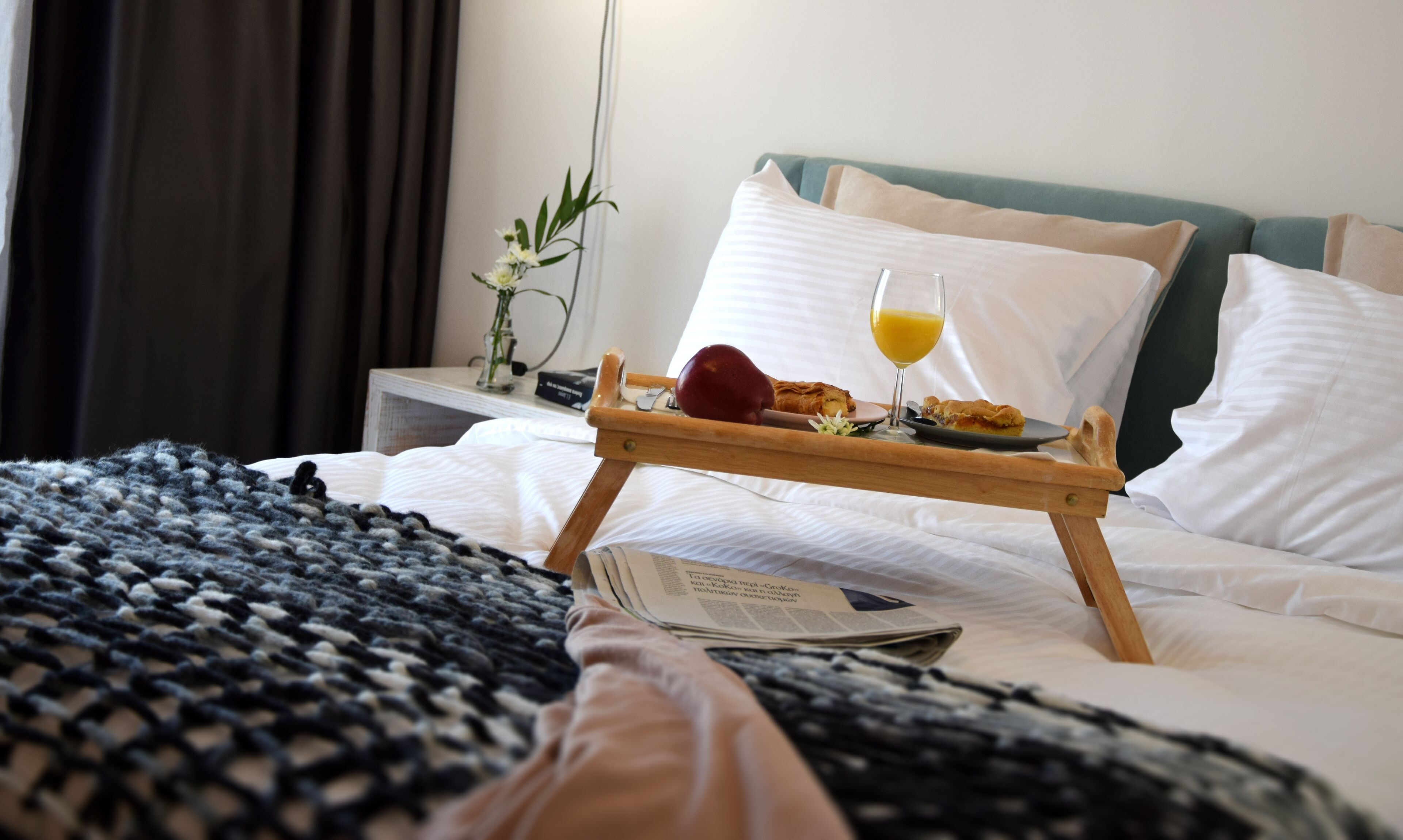 Una cama bien hecha con una bandeja de desayuno, que incluye una manzana roja, un vaso de jugo de naranja y un pastel. Hay un florero con flores en una mesita de noche y un periódico sobre la cama.
