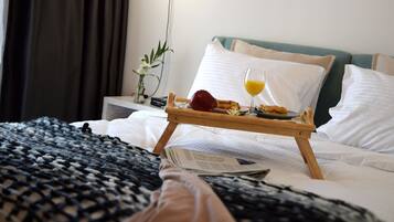 Una cama bien hecha con una bandeja de desayuno, que incluye una manzana roja, un vaso de jugo de naranja y un pastel. Hay un florero con flores en una mesita de noche y un periódico sobre la cama.