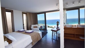 Hotelkamer met een bed, een stoel, een bureau, een groot raam met uitzicht op zee, en een badkamer met een wastafel en spiegel.