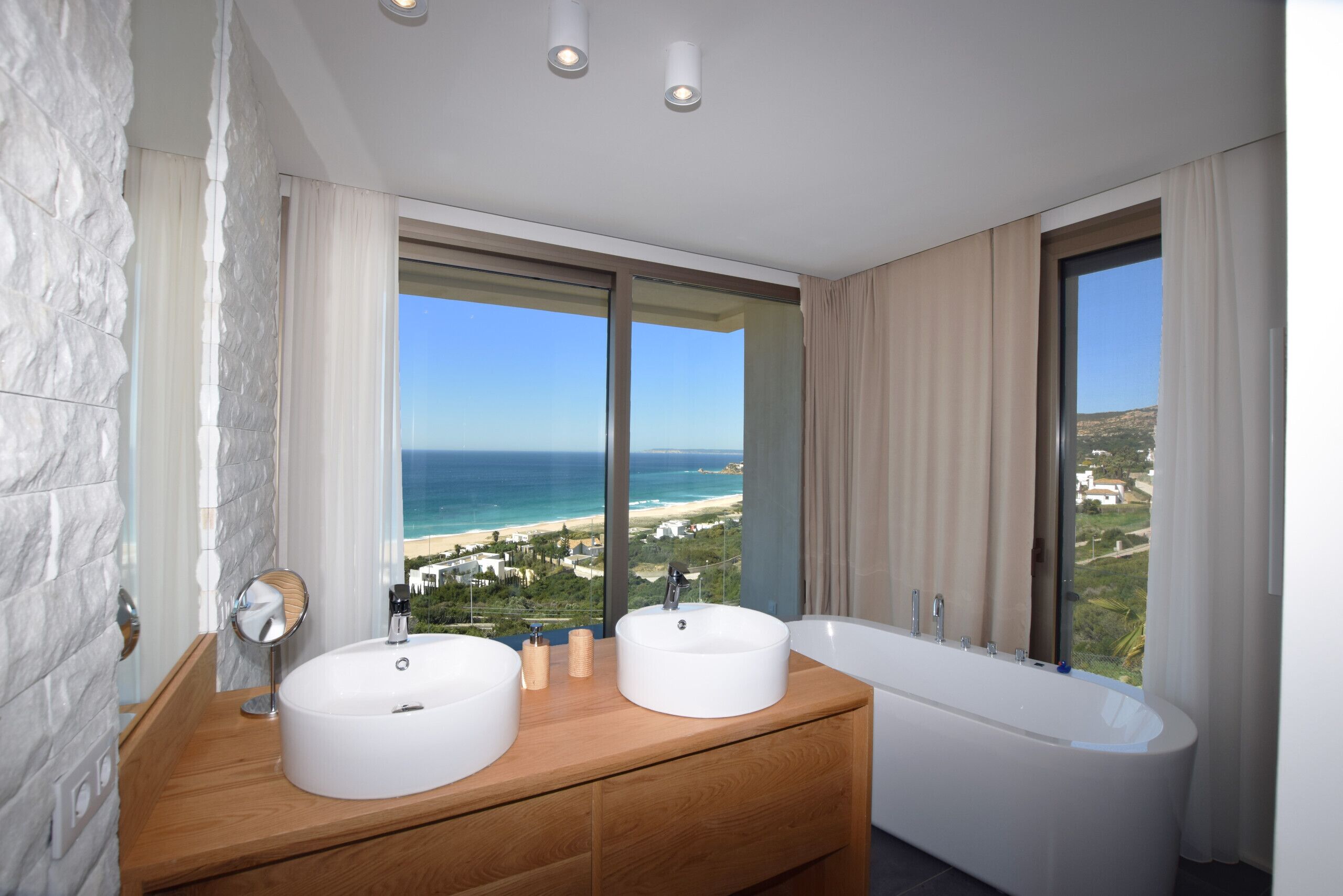 Un baño con dos lavamanos, una bañera y un amplio ventanal con vistas a la playa.