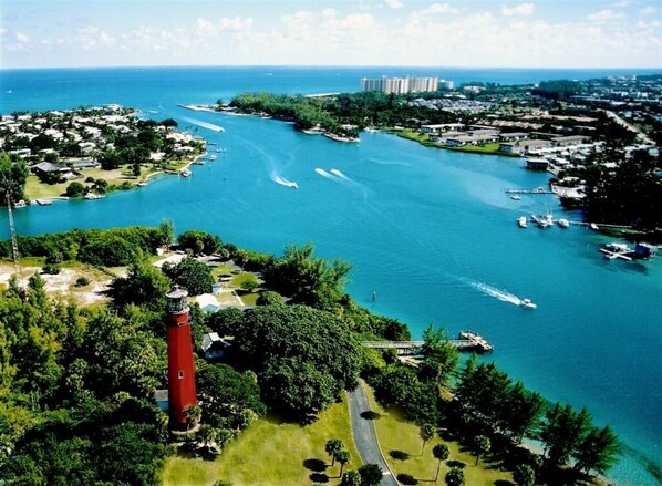 An aerial view of a coastal area with a prominent red lighthouse, a marina with boats, and residential buildings in the background. - BEACHY BUNGALOW. Within two miles of beaches/boat ramps. Book your WINTER stay! (Jupiter)