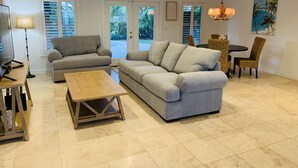 A living room with a grey sofa, a wooden coffee table, a dining table with wicker chairs, and a painting of a beach scene. - BEACHY BUNGALOW. Within two miles of beaches/boat ramps. Book your WINTER stay! (Jupiter)