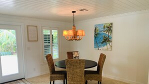 A dining area with a round table and four wicker chairs, a chandelier, a painting of a beach scene, and a door leading to a garden. - BEACHY BUNGALOW. Within two miles of beaches/boat ramps. Book your WINTER stay! (Jupiter)