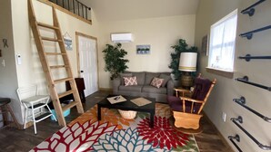 A living room with a sofa, coffee table, and a patterned rug. There is a ladder, a chair, and a lamp. - Grandma's cabin nestled in a little woodland village – playground, pond and more (Winslow)