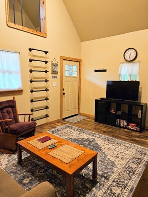 A living room with a wooden coffee table, a television on a stand, a patterned rug, a chair, and a wall-mounted rack. - Grandma's cabin nestled in a little woodland village – playground, pond and more (Winslow)
