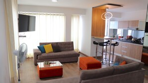 A modern living room with a sofa, ottoman, and a coffee table. There is a kitchen area with bar stools and a TV mounted on the wall. - Residencial Cafferata (Lima)