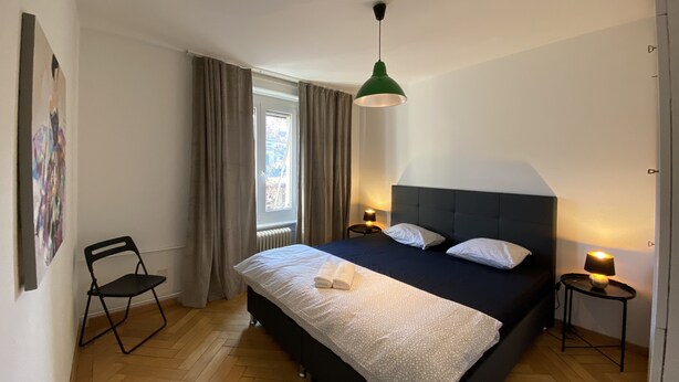 Un dormitorio con cama, una silla, ventana con cortinas, una lámpara colgante verde y dos lámparas de noche.