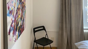 Una habitación con un cuadro en la pared, una silla, un ventanal con cortinas y un radiador.
