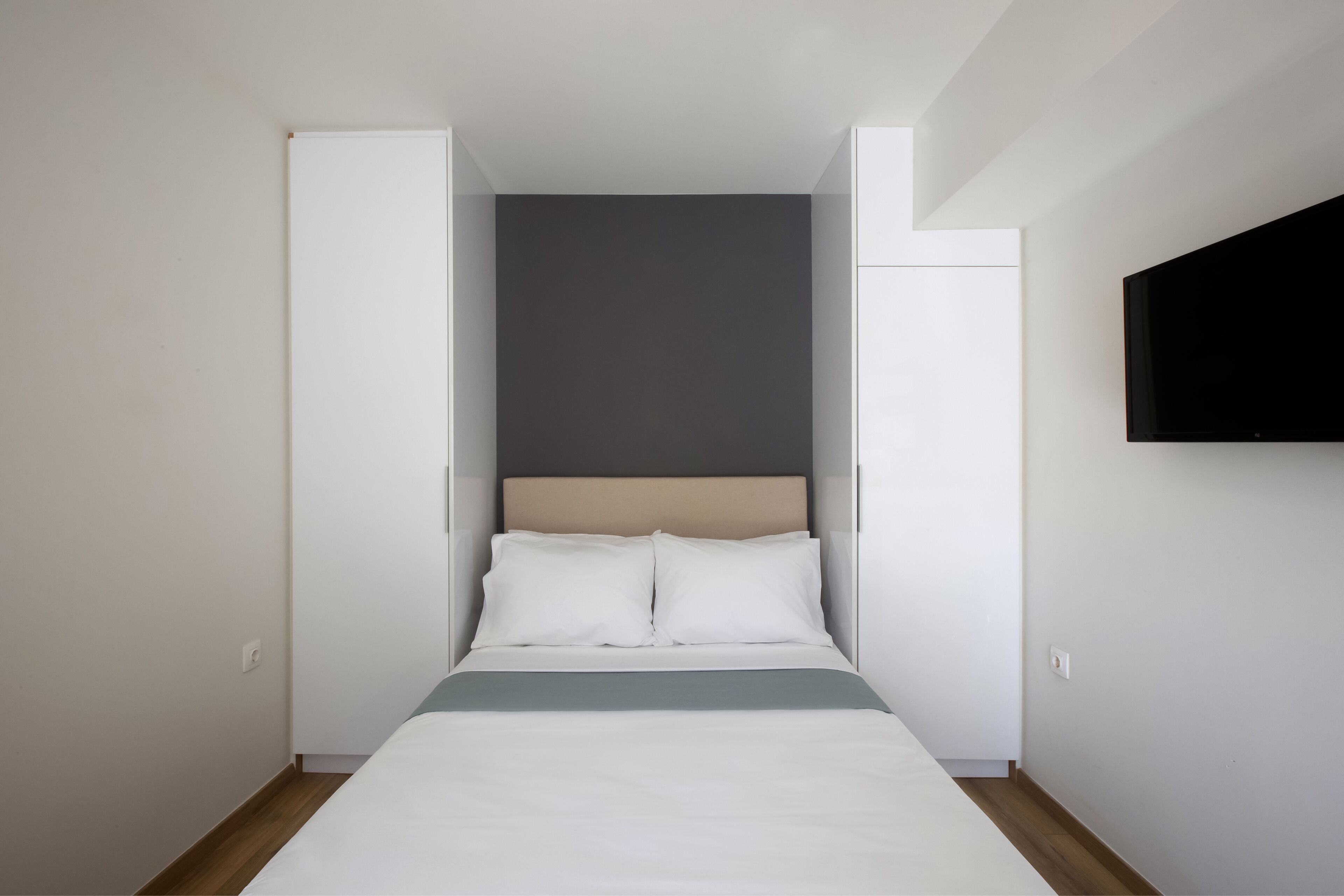 A minimalist bedroom with a bed, a grey headboard, and a TV mounted on the wall.