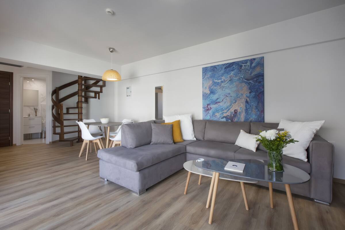 Un salon moderne avec un canapé d’angle gris, une table basse en verre et un coin repas avec des chaises blanches.
