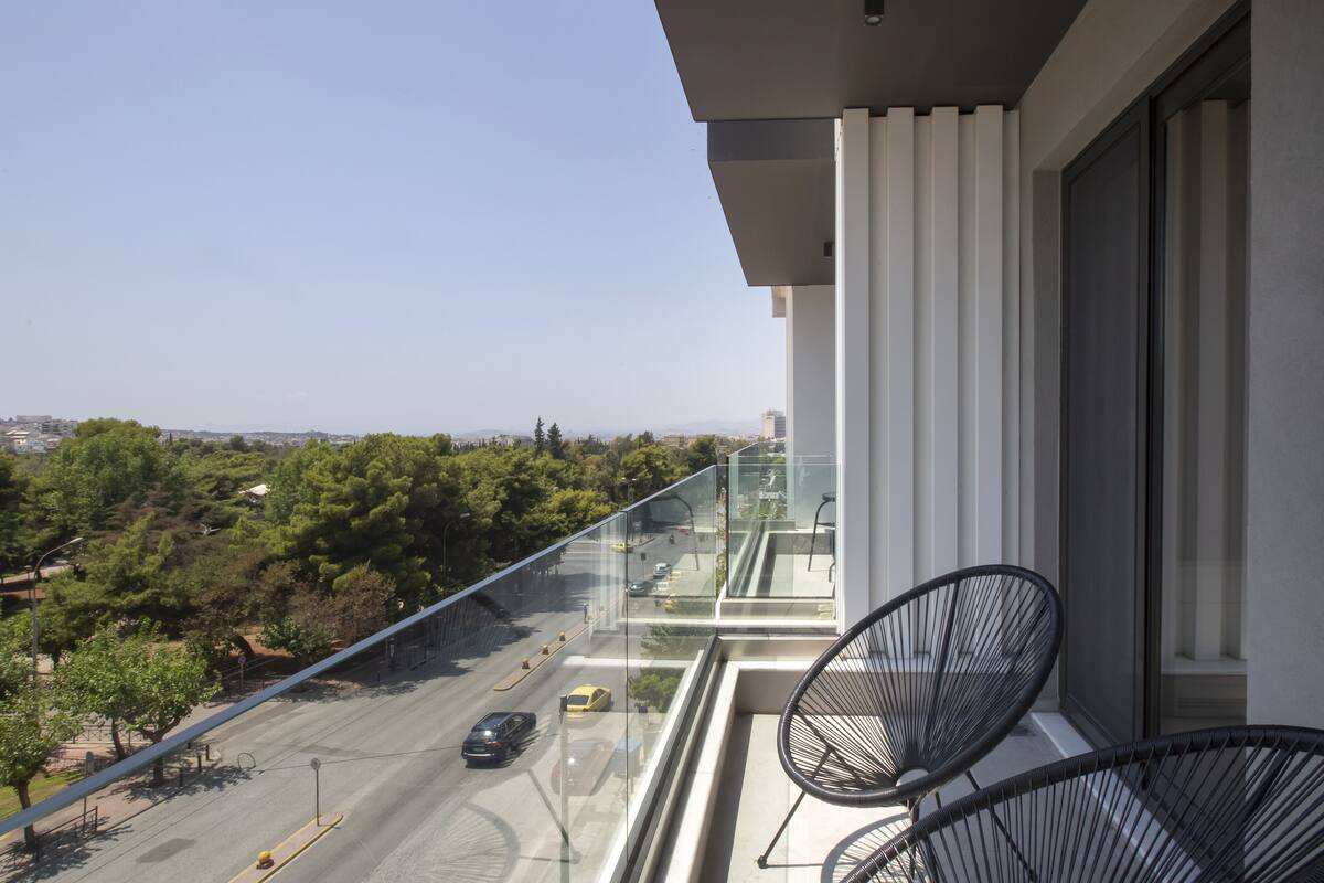 Un balcon offrant une vue dégagée sur une route et la verdure, avec deux chaises noires et une balustrade en verre.